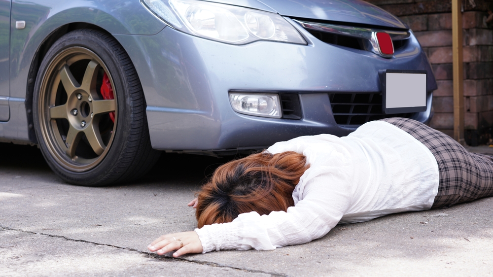 Pedestrian lying on the ground in front of a car after a collision, indicating a possible parking lot accident.
