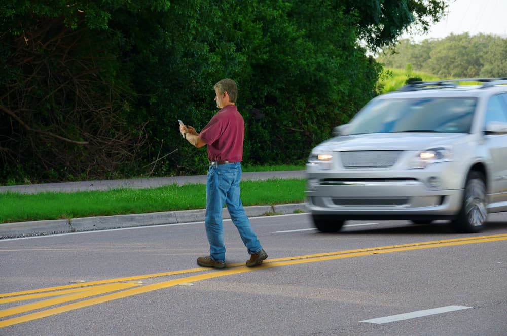 A person wearing a red shirt and blue jeans crosses the street while looking at their mobile phone, with a silver SUV approaching from the right. The background features lush green foliage and a sidewalk, emphasizing urban pedestrian safety and distracted walking risks.